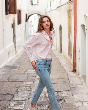 A person wears MERSEA’s Murphy Mini Boot Cut Jean in Sea Spray—light blue, 4-way stretch denim—paired with a white top and beige slide sandals, standing on a white floor before a beige fabric backdrop. Their face is not visible.