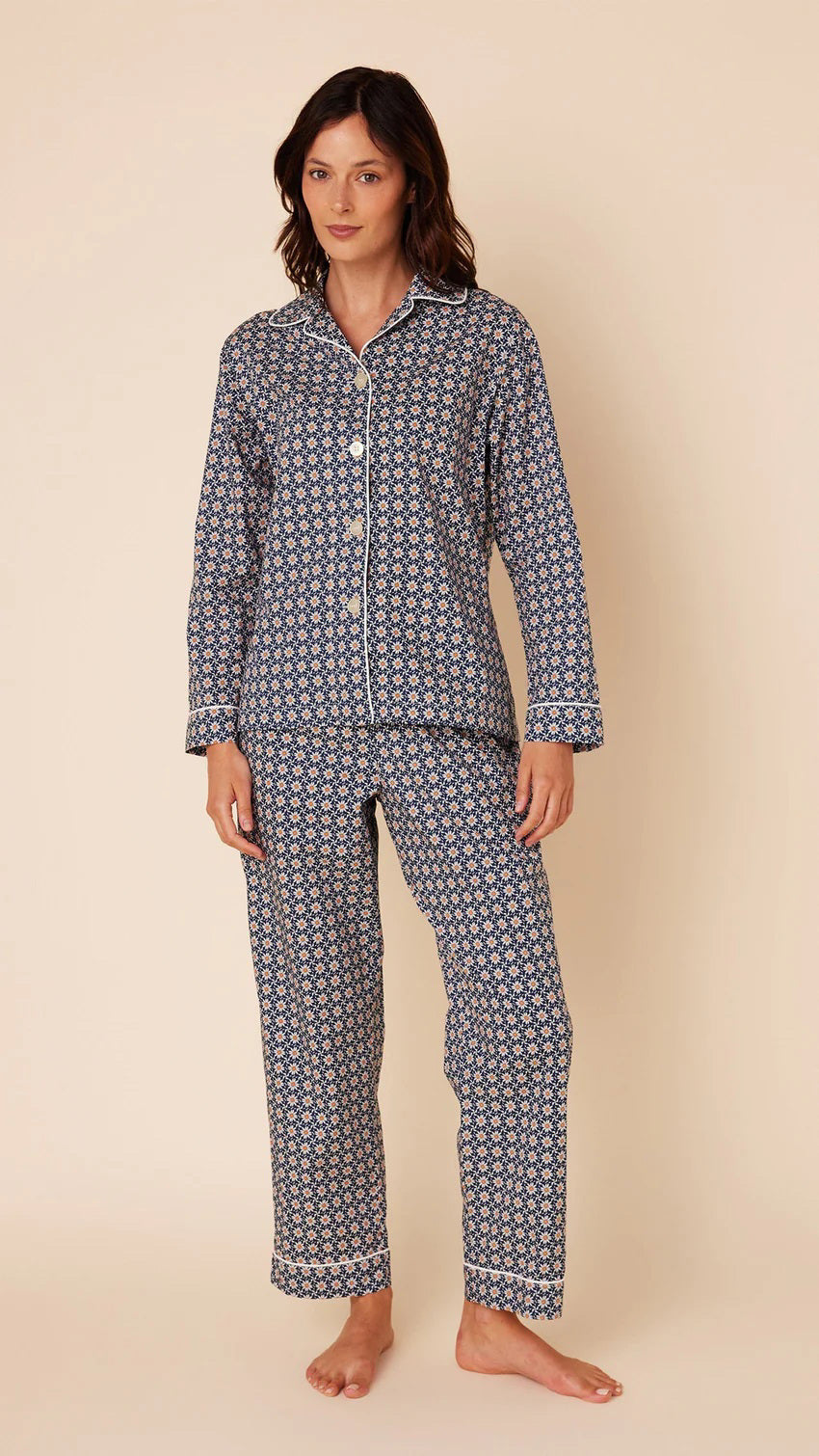 A person stands against a beige background wearing the CAT&#39;S PAJAMAS Newport Daisy Luxe Pima Cotton Pajamas by CAT&#39;S PAJAMAS. The navy blue pajama top features a white geometric pattern, long sleeves, a collar, and white buttons down the front. The matching full-length pajama pants complete the set. The person is barefoot.