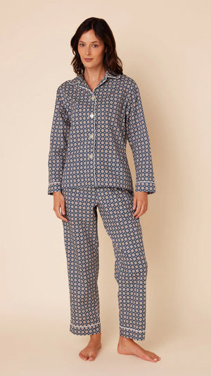 A person stands against a beige background wearing the CAT'S PAJAMAS Newport Daisy Luxe Pima Cotton Pajamas by CAT'S PAJAMAS. The navy blue pajama top features a white geometric pattern, long sleeves, a collar, and white buttons down the front. The matching full-length pajama pants complete the set. The person is barefoot.