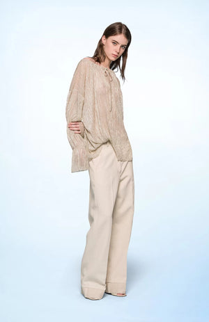 A person with shoulder-length hair stands against a light blue background, wearing the SEVENTY VENEZIA LUREX JERSEY BLOUSE by SEVENTY, which is a shimmery beige pleated blouse with long sleeves, paired with wide-leg beige trousers. The person gazes forward with a relaxed posture, one hand in their pocket.