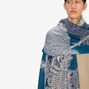 A blonde woman in a black coat stands against a white background, wearing the INOUI EDITIONS "Owls on a Branch" wool scarf in blue by INOUI, draped over her neck and one shoulder with intricate white owl designs.