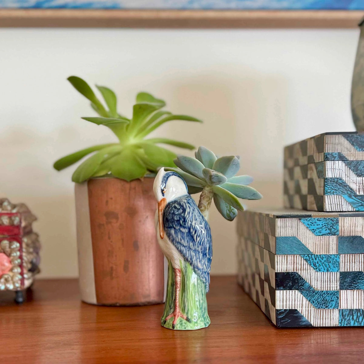 The QUAIL CERAMICS - HERON BUD VASE by QUAIL DESIGNS features a hand painted stoneware heron with blue and white feathers, orange beak, and green legs perched on a green base, shown against a plain white background.