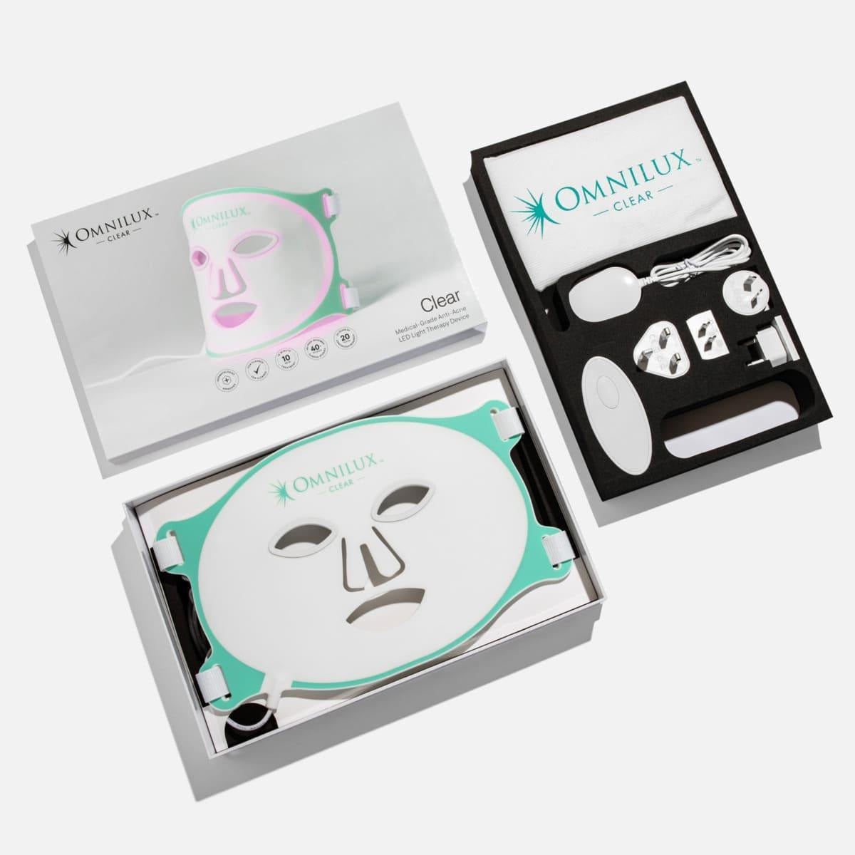The OMNILUX - CLEAR MASK is a white and teal LED light therapy mask for acne with eye, nose, and mouth cutouts. It emits a pink glow and has two side adjustable straps. Dermatologist-recommended, it sits against a plain background.