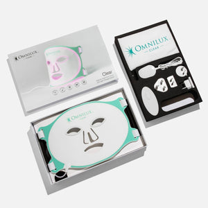 The OMNILUX - CLEAR MASK is a white and teal LED light therapy mask for acne with eye, nose, and mouth cutouts. It emits a pink glow and has two side adjustable straps. Dermatologist-recommended, it sits against a plain background.
