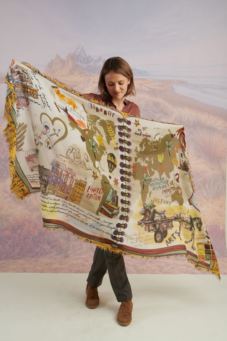 A woman with wavy blonde hair smiles while looking down. She wears a loose, light lavender blouse, dark pants, and the STORIATIPIC EUGENIE WOOL SCARF. Behind her is a scenic landscape of mountains, grass, and water.