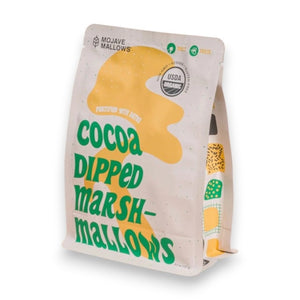 A white upright bag of MOJAVE MALLOWS - COCOA DIPPED MARSHMALLOWS by MOJAVE MALLOWS features green and yellow text, a bright yellow graphic, and a USDA organic label on the front, highlighting this delicious handmade treat. Each marshmallow is infused with organic vanilla and coated in rich dark cocoa powder for an irresistible snack.