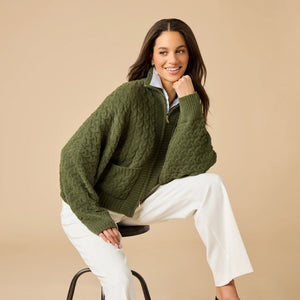 A woman wearing a cream-colored, plush SOFTIES - CABLE KNIT MARSHMALLOW FUNNEL NECK DOUBLE ZIP JACKET with a funnel neck and double zipper, paired with blue jeans.