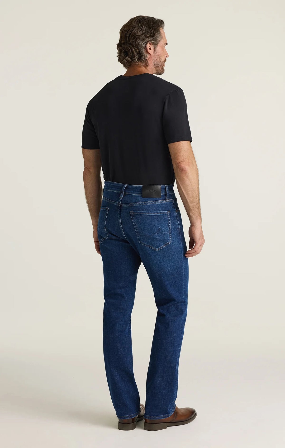 A person is shown from the back wearing a black shirt and the 34 HERITAGE - CASH BOOTCUT JEANS IN DARK BRUSHED VINTAGE by 34 HERITAGE. The vintage denim features back pockets and a black leather patch on the waistband, with the wearer’s arms visible.