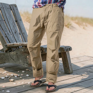 Wearing RELWEN's Flyweight Flex Chino in British Khaki, a person stands on a beachside boardwalk near a weathered bench, with flip-flops and a plaid shirt, seashells and sand in the background.