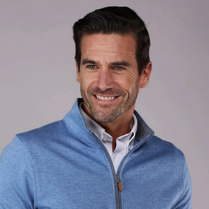 A man with short dark hair and a beard smiles while wearing the COLLARS AND CO - HEATHER CLOUD COTTON 1/4 ZIP SWEATER IN MEDIUM BLUE WITH GREY TRIM over a white collared shirt, standing against a plain gray background.