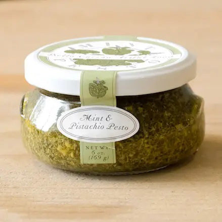 A small glass jar filled with green pesto labeled "BELLA CUCINA - MINT PISTACHIO PESTO" is shown. The white lid has a green design, and a seal runs from the lid to the base, marked with the same label. Made with olive oil and fresh mint leaves, the jar sits on a reflective surface against a plain background.