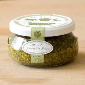 A small glass jar filled with green pesto labeled "BELLA CUCINA - MINT PISTACHIO PESTO" is shown. The white lid has a green design, and a seal runs from the lid to the base, marked with the same label. Made with olive oil and fresh mint leaves, the jar sits on a reflective surface against a plain background.
