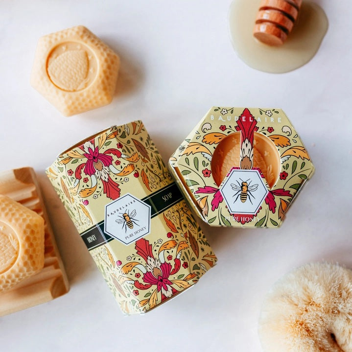 A yellow floral BAUDELAIRE box labeled “PURE HONEY SOAP” is displayed beside a stack of four light orange, honeycomb-shaped triple-milled soaps from the BAUDELAIRE PURE HONEY SOAP 1.4 OZ QUARTET, each featuring a textured surface.