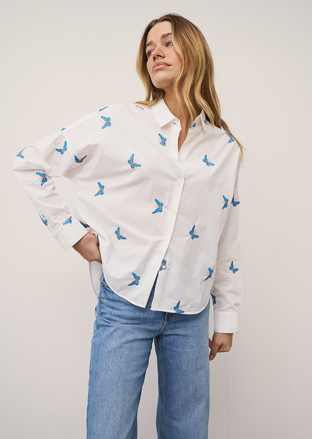 A woman with long, straight hair stands against a plain background wearing the 0039 ITALY LUCY BLOUSE WITH EMBROIDERED BUTTERFLIES, light blue wide-leg jeans, and silver sandals. She is looking slightly to the side.