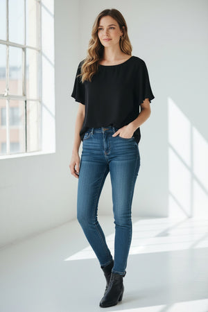 A woman with long, wavy blonde hair models the MEIMEIJ FLUTTER SLEEVE BLOUSE, paired with bold black floral print pants. The refined look is effortless and perfect for washable silk styles, set against a light textured background.