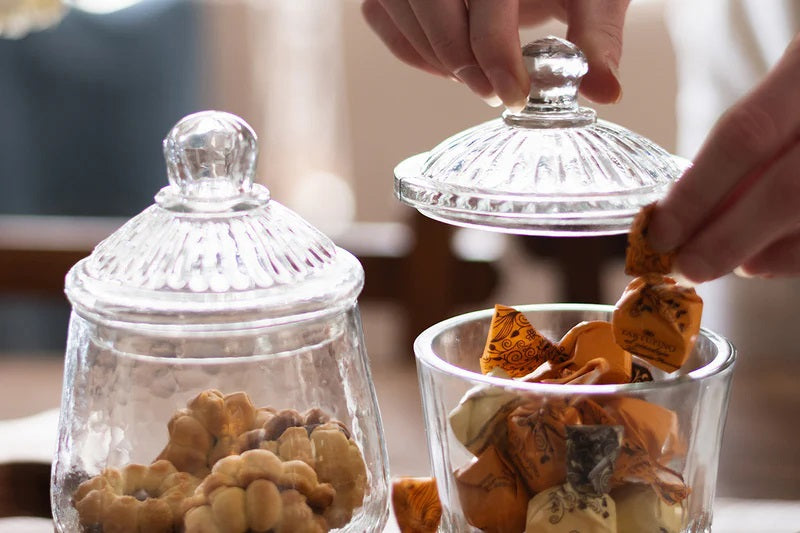 Two ETUHOME Bistro Glass Bon Bon Jars with ribbed lids sit on a wooden surface, one filled with yellow candies and the other nearly empty. Wrapped sweets are scattered nearby, creating a charming ETUHOME display.