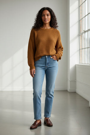 A woman with wavy blonde hair wears a BASE MILANO - CREWNECK COTTON BLEND SWEATER in a relaxed fit and blue jeans, standing against a plain light background as she looks down and gently pulls up one sleeve.