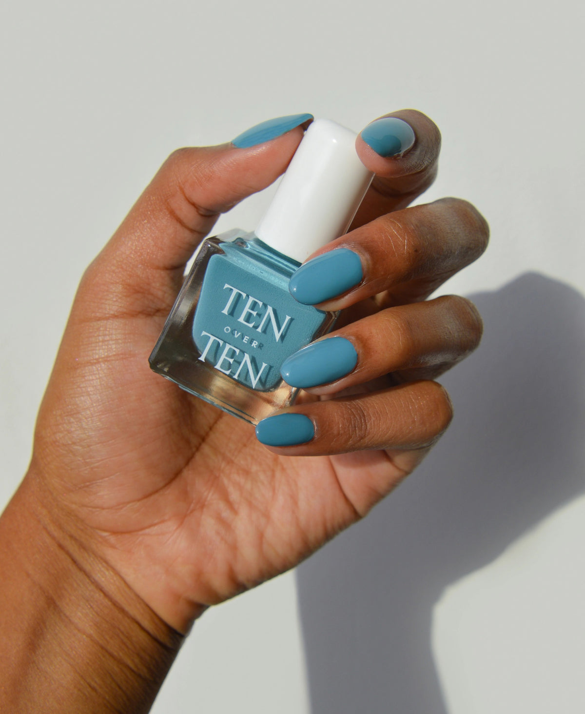 A glass bottle of TENOVERTEN - NAIL POLISH from the brand TEN OVER TEN, featuring a white cap and a dusty pink nail color. The front label displays "TEN OVER TEN" in white uppercase letters. This vegan, eight-free formula ensures both beauty and peace of mind. The background is plain and light-colored.