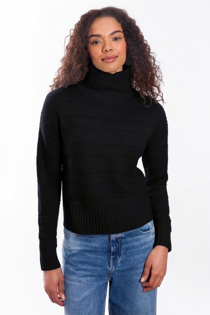 A person with curly hair, dressed in the & ISLA - TEXTURED MOCK NECK PULLOVER IN BLACK by & ISLA and blue jeans, stands against a plain white background facing the camera with a slight smile.
