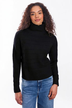 A person with curly hair, dressed in the & ISLA - TEXTURED MOCK NECK PULLOVER IN BLACK by & ISLA and blue jeans, stands against a plain white background facing the camera with a slight smile.