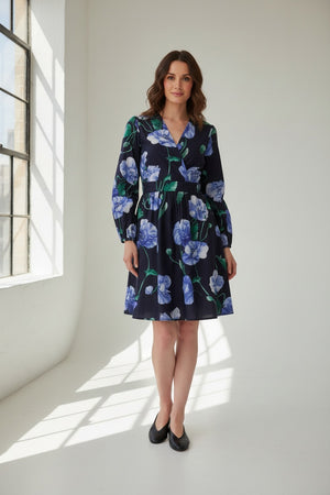 A woman wearing the LAVI Cetara Dress in Blue Poppies by LAVI—an Italian cotton, navy blue, long-sleeve dress featuring a green and blue botanical print—paired with beige sandals, stands against a plain white background.