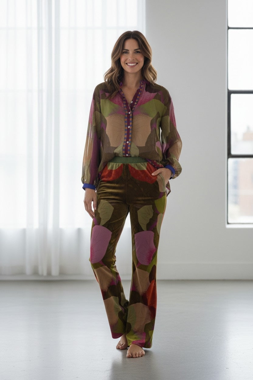 A woman stands barefoot in a bright room, smiling, wearing the FRANCO FERRARI - KANADA VELVET FLARE TROUSER paired with a sheer, patterned blouse; both pieces feature earthy tones with pink, purple, and green abstract shapes.