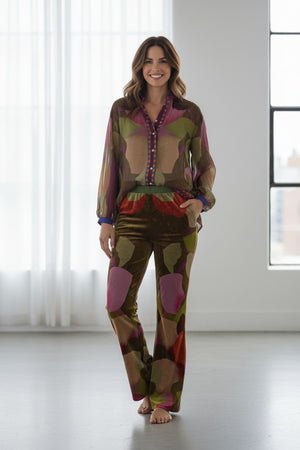 A woman stands barefoot in a bright room, smiling, wearing the FRANCO FERRARI - KANADA VELVET FLARE TROUSER paired with a sheer, patterned blouse; both pieces feature earthy tones with pink, purple, and green abstract shapes.