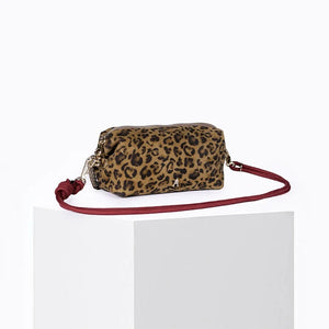 The CRAIE STUDIO - NUAGE LEO SEPIA MINI BAG, featuring a leopard print design, gold zipper, and an extended red strap, is showcased on a white pedestal against a simple backdrop.