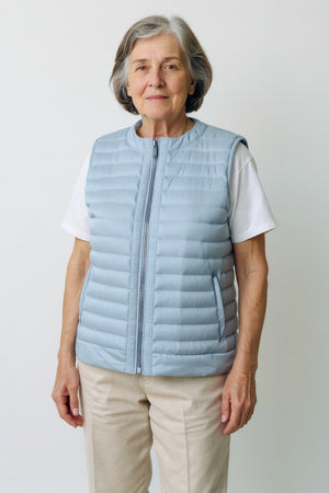 An older woman with short gray hair wears the SAVE THE DUCK - MARA PUFFER VEST over a white t-shirt and beige pants, standing against a light background—perfect for spring–summer layering from SAVE THE DUCK.