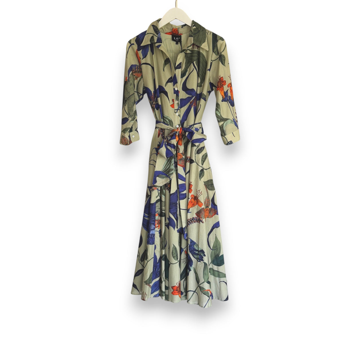 Against a plain background, someone models the LAVI COUTURE SPRITZ DRESS by LAVI, featuring green and pink botanical prints. This long Italian garden dress has long sleeves, a tied belt, and pockets, all crafted from breathable cotton. Their pink shoes complete the look elegantly.