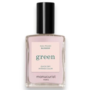 A hand with pale pink polished nails holds a bottle of MANUCURIST INC GREEN™ Nail Polish in Blossom against a gray knit sweater background.