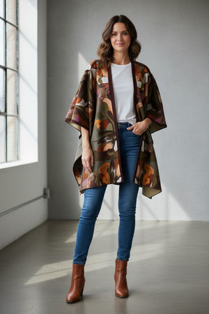 A woman stands indoors in front of large windows with natural light streaming in, wearing jeans, brown ankle boots, a green top, and the FRANCO FERRARI - WOOL CAPE by FRANCO FERRARI—a stylish piece perfect for lightweight layering.