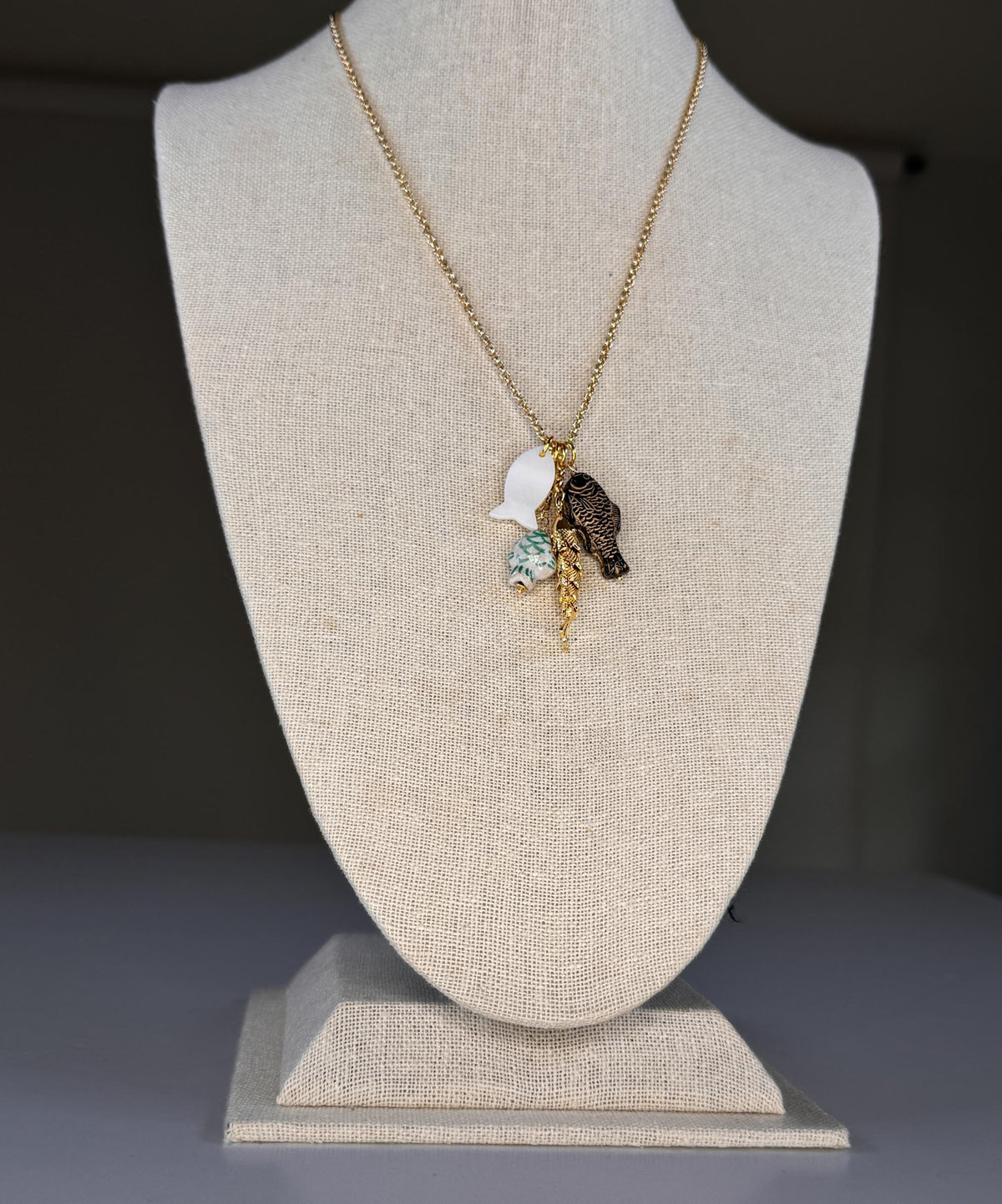 The SANDRINE FRANCE STUDIO - CORDELIA NECKLACE features handmade gold chain with four pendants—a black fish, a gold fish, white sea glass-like piece, and a ceramic School of Fish—beautifully displayed on a plain white surface.