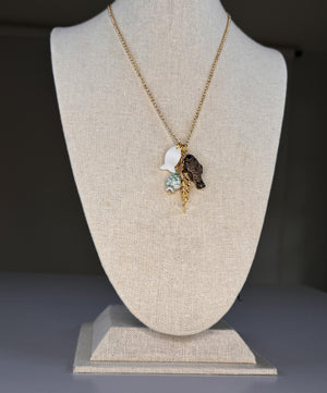 The SANDRINE FRANCE STUDIO - CORDELIA NECKLACE features handmade gold chain with four pendants—a black fish, a gold fish, white sea glass-like piece, and a ceramic School of Fish—beautifully displayed on a plain white surface.