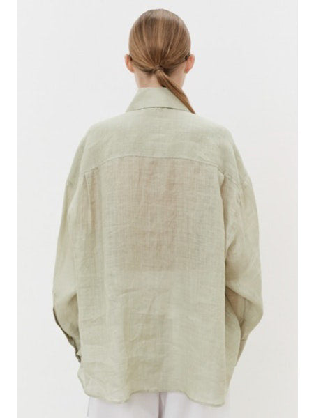 A person with brown hair in a low ponytail faces away, wearing an oversized MEIMEIJ - LINEN SHIRT. Against a plain white background, breathable linen pants are visible at the bottom.