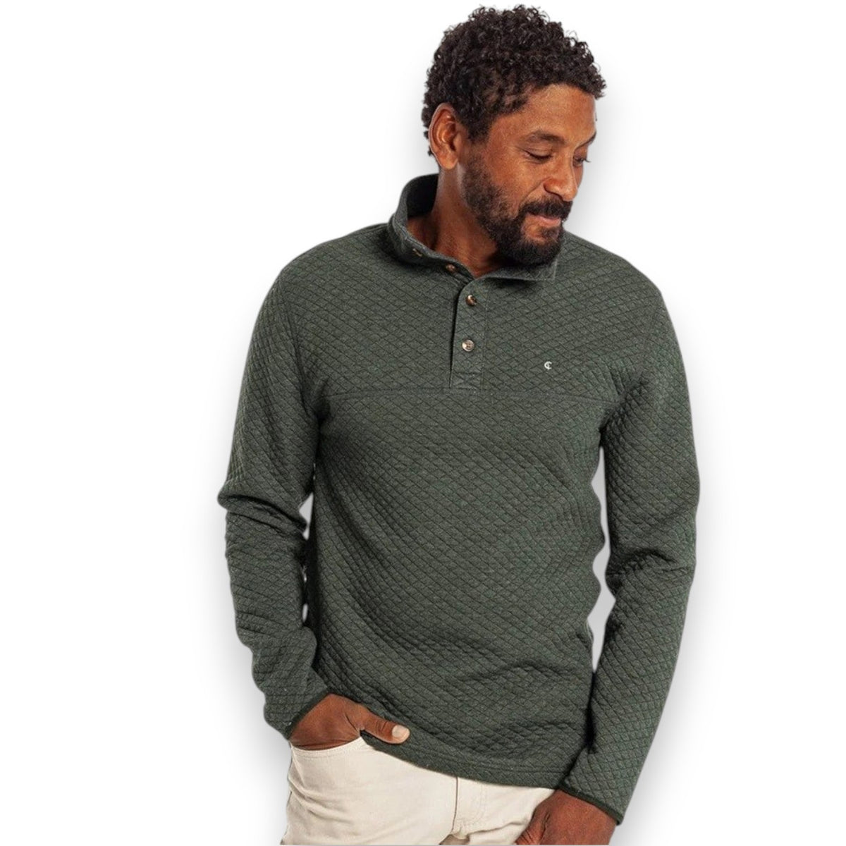Wearing the CRIQUET Quilted Pullover in Heather Ivy, a person stands by a stone wall in khaki pants, holding a patterned mug and smiling, with greenery in the background—highlighting this classic-fit all-season layer from CRIQUET.