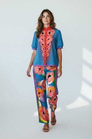 A woman wears the FRANCO FERRARI COTTON PRINT TOP, featuring bold pink and orange floral patterns, puffed sleeves, and a matching multicolored set. The Italian-made piece radiates vibrant style in a brightly lit room.