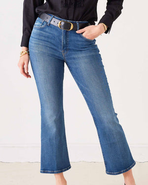A woman stands against a plain white background wearing sunglasses, a black long-sleeve blouse, black heels, and holding a black handbag. She completes her look with the MERSEA Murphy Mini Boot Jean in Tide from MERSEA—blue, cropped organic cotton jeans designed for a gap-free waist.