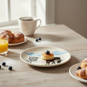 A bright breakfast nook showcases the FEARRINGTON SIGNATURE COLLECTION - BELTIE DISH BY VIETRI, holding a small pancake with a blueberry, surrounded by coffee, orange juice, croissants, and scattered blueberries.