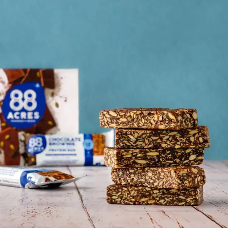 A 88 Acres Dark Chocolate Brownie High Protein Bar wrapper rests on a white wooden surface. The predominantly white and blue wrapper features the brand name &quot;88 Acres&quot; alongside the product name &quot;Rich &#39;n&#39; Fudgy Dark Chocolate Brownie Pumpkin Seed High Protein Bar.&quot; Text highlights that it is nut-free, vegan, and gluten-free.