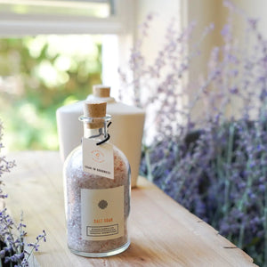 A cork-stopped glass jar of ETHICS SUPPLY CO's pink "Soak in Goodness" Bubbly Bath Salt Soak sits on a white surface, accompanied by a natural sea sponge. Infused with essential oils, this cruelty-free treat promises relaxation and features a tag reading "Soak in goodness.