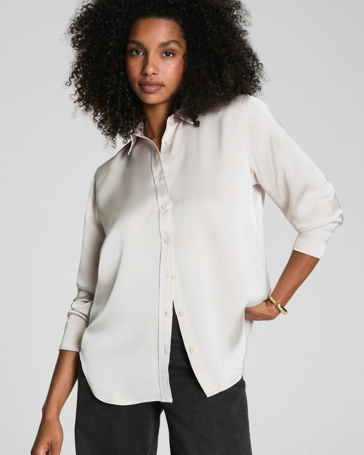 A woman with curly hair wears the SPANX Silky Smooth Button Down Shirt in a relaxed tailored fit and black pants, turning slightly as she looks over her shoulder against a plain gray background.