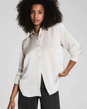A woman with curly hair wears the SPANX Silky Smooth Button Down Shirt in a relaxed tailored fit and black pants, turning slightly as she looks over her shoulder against a plain gray background.