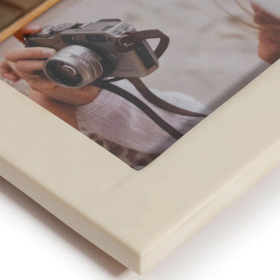 A person in a white hat, looking down and holding a camera, is pictured in the ZODAX WHITE HORN AND IVORY RESIN FRAME - 4X6, which features a stylish design crafted with natural horn and ivory resin inlay.