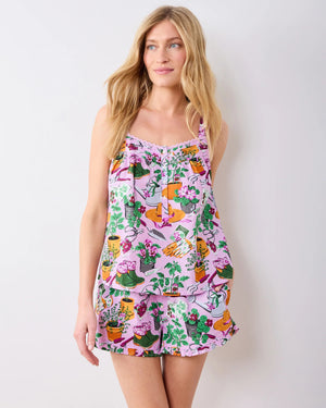 Wearing the PRINTFRESH Green Thumb Back to Bed Pajama Short Set—a vibrant, printed sleepwear ensemble from PRINTFRESH—a woman stands smiling in a lush, green garden, glancing to the side.