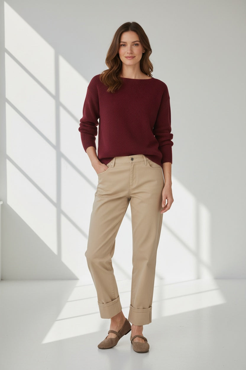 A woman stands in a bright room wearing a maroon sweater, brown shoes, and the BRAX - MADISON FIVE POCKET STRAIGHT FIT pants in Almond by BRAX, featuring a subtle cotton satin finish. She has wavy hair and is smiling slightly as sunlight casts gentle shadows on the white walls and floor.
