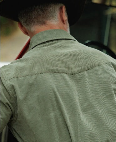 A smiling man with a gray beard, wearing the CRIQUET Long Sleeve Corduroy Pearl Snap Shirt in Olive and a brown cowboy hat, stands by the open door of a red and white vehicle.