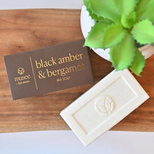 A rectangular white moisturizing bar soap featuring an embossed leaf logo sits beneath black packaging labeled in gold with “MUSEE - BLACK AMBER & BERGAMOT BAR SOAP,” showcasing the luxurious blend of MUSEE’s Black Amber and refreshing bergamot.
