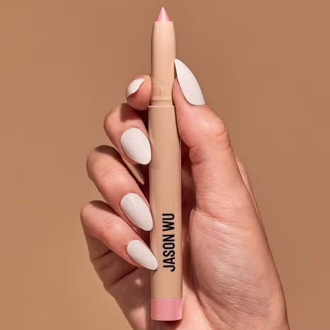 Close-up of a hand with white painted nails holding a sparkle pearl colored eyeshadow pencil with a pink tip, branded with &quot;JASON WU BEAUTY&quot; in black text against a solid neutral beige background.