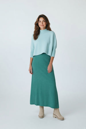 A person wears a light blue cropped turtleneck sweater with a BRITON COURT - TIFFANY MIDI SKIRT IN EMERALD STRIPE and beige ankle boots.